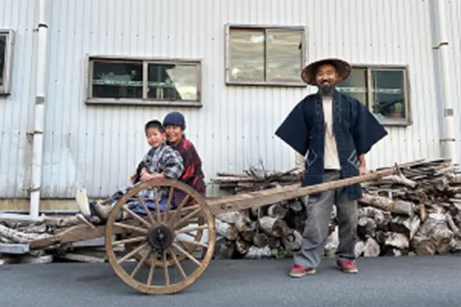 建物の前で、菅笠をかぶった男性と、木製の荷車に乗った親子が並んでいる様子。