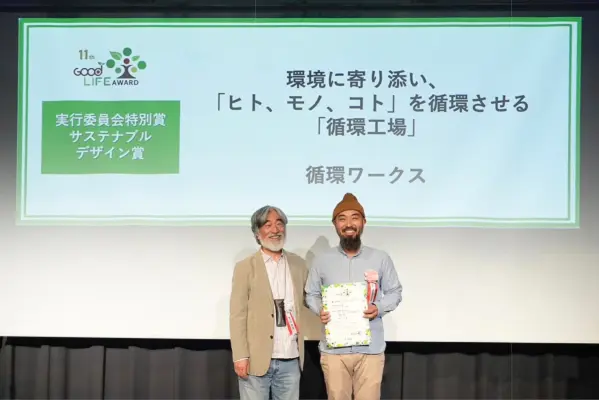 表彰式で、ニット帽をかぶった男性が賞状を手に、年配の男性と並んで立っている。