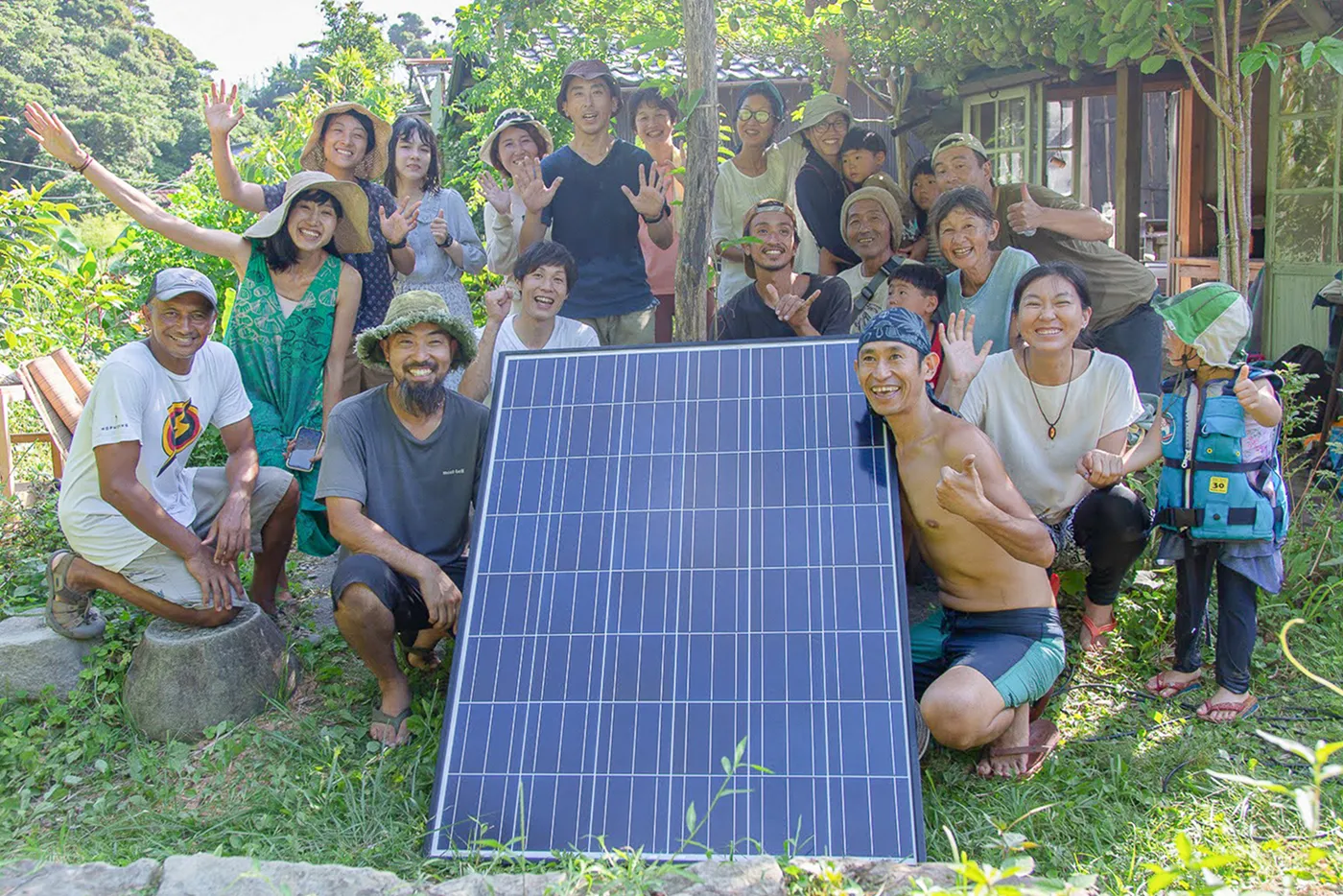 緑豊かな屋外で、大きなソーラーパネルを囲んで大勢の人が集まり、笑顔で記念撮影をしている。