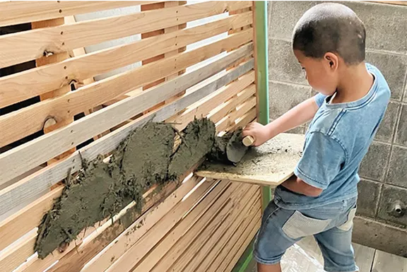 青いTシャツの子供が、木の板でできた壁にコテを使って土のようなものを塗りつけている。