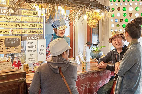 カフェのカウンターで、店員の女性と客の男女3人が話している。皆が帽子をかぶっている。