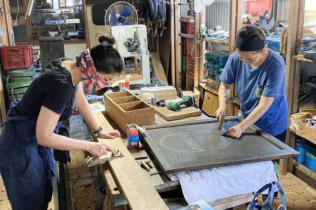 工房のような場所で、二人の女性が木材を加工する作業をしています。