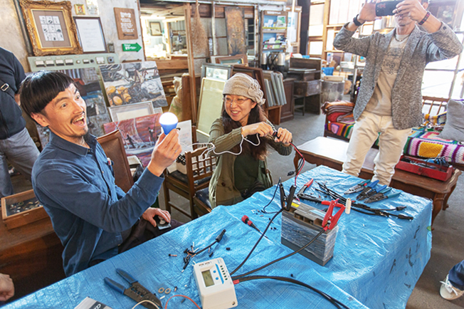 青いシートを敷いた机の上で、バッテリーに繋いだ電球が点灯。電球を手にした男性が嬉しそうな顔をしています。