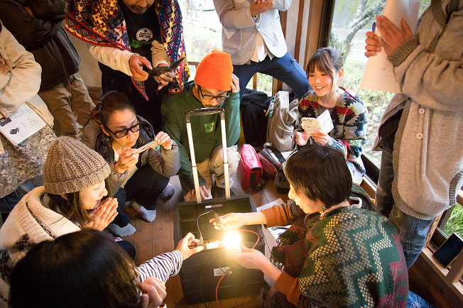 たくさんの人が輪になり、床に置かれた黒い箱から光る電球を、興味深そうに覗き込んでいる様子です。