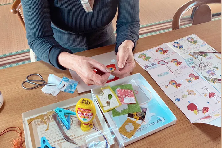 和室の机で、女性が手芸をしている様子。机にはハサミや接着剤、布などの道具が広げられています。