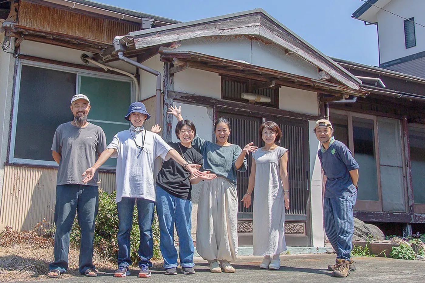 晴れた日に、古い日本家屋の前で6人の男女が横一列に並んで記念撮影をしている。