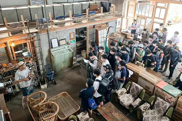 倉庫のような場所で、一人の男性が大勢の生徒たちに何かを説明している。生徒たちは男性を取り囲んで聞いている。