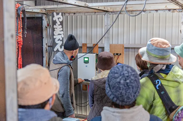 ニット帽の男性が、壁に取り付けられた白い機械を指さしながら、周りの人たちに説明している場面。