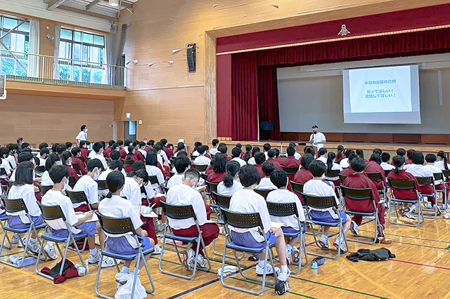 体育館で、たくさんの生徒たちが椅子に座っている。ステージ上のスクリーンを見ながら、男性の話を聞いている様子。