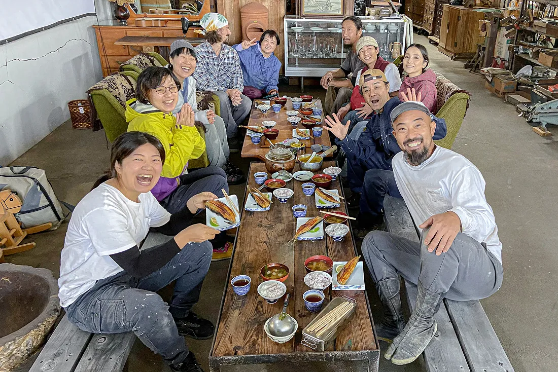 作業場のような広い空間で、10人の男女が長いテーブルを囲んで食事をしている。皆が笑顔で写っている。