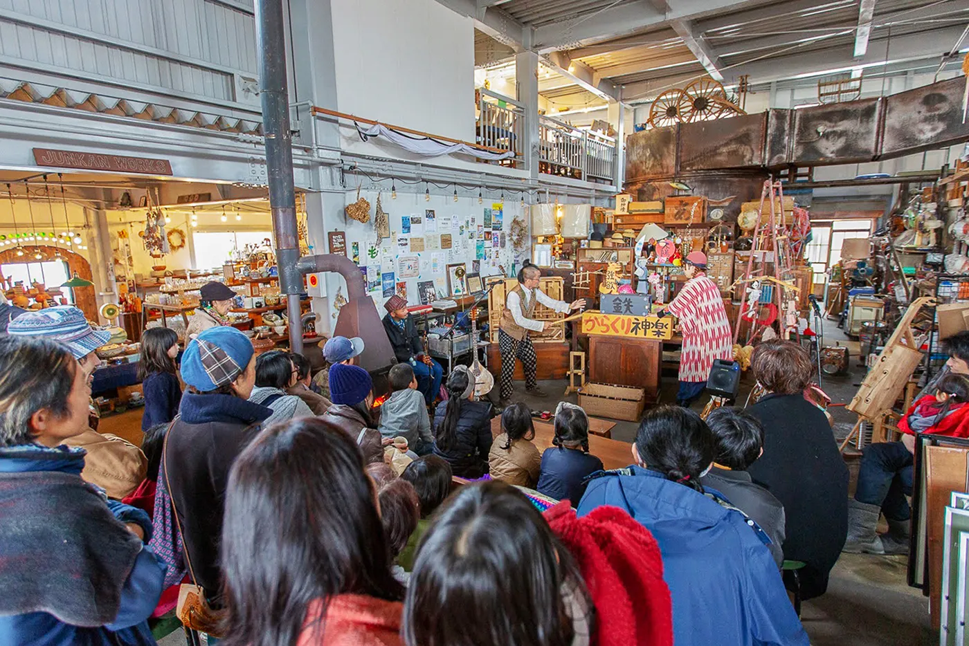 古道具がたくさん置かれた広い室内で、大勢の観客が男性2人のパフォーマンスを座って見ている様子。